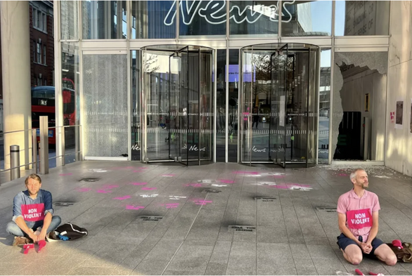 Protesters from Extinction Rebellion sit calmly outside News UK HQ in London, awaiting arrest on the hottest day on record, 2022 after smashing windows in peaceful direct action. The protesters have patches on their chests which say 'non violent'. Two windows of the glass front of the building are shattered. Spray paint stencils  "tell the truth" can be seen on the floor.