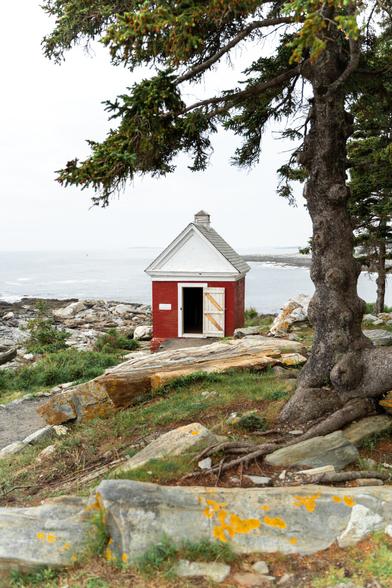 A digital photograph of a small red brick building with white trim and a shingle roof. The building is built on the rocks at Pemaquid Point Lighthouse and may have been used for storage of lamp oil back in the day though I’m not positive. In the foreground there are slabs of rock with bright yellow lichen growing on them and a gnarled spruce tree in the righthand side of the frame. In the background the rocks descend down to the shoreline of the gulf of Maine and there is a point of land in the distance. The day is hazy; the horizon line is obscured and the sky appears white.