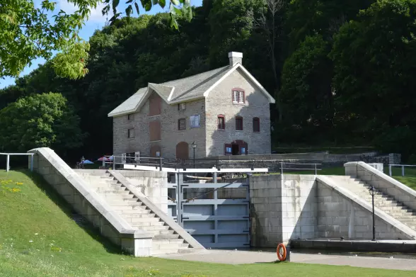 The image depicts a building situated near a canal or waterway. The structure appears to be made of stone and has several windows and a chimney. In front of the building, there are steps leading down to what looks like a lock or a water control system. The surrounding area is green with trees, suggesting a natural setting. There seems to be a pathway or recreation area, likely used for walking or visiting the site.

Image Credits: Wikimedia / Bytownmuseum / CC BY-SA 4.0