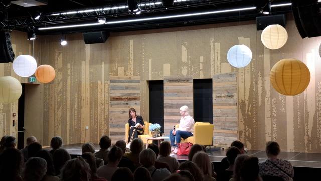 Bad snapshot, looking across the first 10 rows or so, the backs of heads almost in darkness, towards the illuminated stage in the main event room at Dokk1 Aarhus Main Library. Big round paper-shade lamps hang from the ceiling. In front of a wooden LiteratureXchange backdrop, sitting in comfy yellow armchairs:  Rachel Cusk and literature professor Elisabeth Friis. Friis is white, has white hair and is wearing pale pink. Cusk is white, has dark hair and is wearing dark green. She is reading from her book Parade.