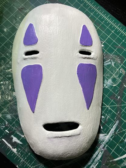 A painted plastic mask sits on a green cutting may. The mask is white with purple triangles above and below the small rectangular eye holes.