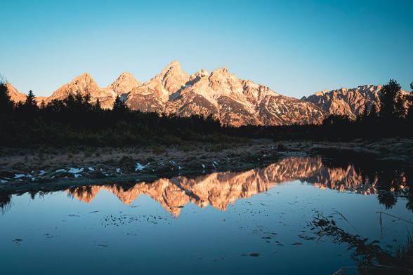 The sun shining directly on sharp peaked mountains mirrored in a still lake with trees lining the shore.