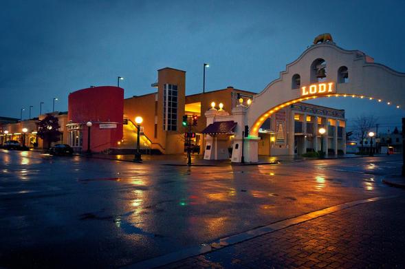 Street lights illuminate puddles of water reflections in the evening hours after a heavy rain storm. A small town's gateway arch and adjacent street buildings are the main focus of the image.