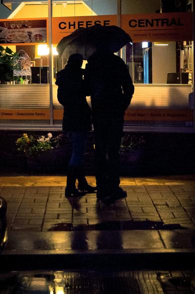silouette of a male and female under an umbrella peering in to the storefront window of a cheese shop closed for the evening. Light reflects on the street and sidewalk off of puddles of water after a rain storm