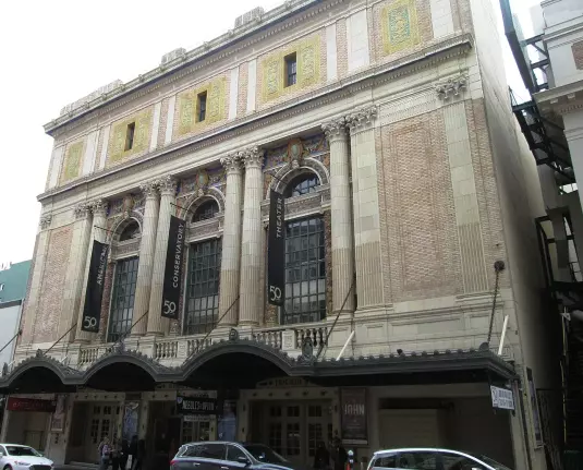The image shows the exterior of a theater building. The architectural style features ornate details, including columns and decorative mosaics. There are banners hanging from the front, indicating events or anniversaries, and a marquee at the entrance. The building appears to be located in an urban area, likely a place known for performances and events.

Image Credits: Wikimedia / Beyond My Ken / CC BY-SA 4.0
