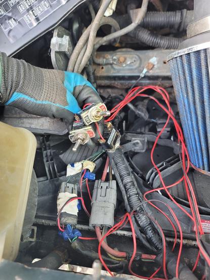 Photo of a gloved hand holding a group of wired switches. They are connected to the inside of an RX-8 engine bay.