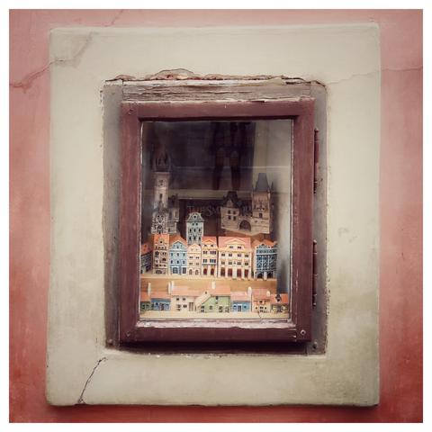 Quadratisches Foto mit weißem Rand. Zu sehen ist ein kleines Fenster in einer rötlichen Hauswand. Im Fenster steht eine kleine Stadt aus Keramikhäusern.