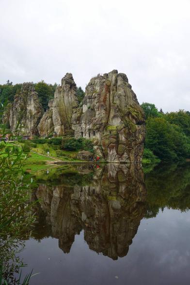 A wide, eye-level shot on an overcast day captures the Externsteine rock formation, a natural monument in North Rhine-Westphalia, Germany. Three imposing, weathered sandstone pillars, richly textured with fissures and patches of moss, rise majestically from a lush green landscape. The largest rock, on the right, features carved openings resembling windows or caves. To the left, a smaller pillar stands slightly behind the main formation, with a third, even smaller one partially visible further left, nestled amongst trees.
The rocks are reflected with striking clarity in the still, dark water of a lake in the foreground, creating a symmetrical visual effect. The banks of the lake are verdant, with green grass and foliage. A small pathway or boardwalk with a wooden fence is visible at the base of the rocks, suggesting accessibility for visitors. The overall scene conveys a sense of natural grandeur and historical significance.