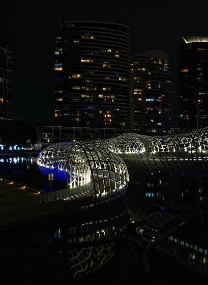 Webb Bridge is a striking pedestrian bridge located in Melbourne, Australia, known for its unique and artistic design. Completed in 2003, it was a collaborative project between artist Robert Owen and architects Denton Corker Marshall. The bridge repurposes the structure of an old rail bridge and features a woven, cocoon-like steel mesh that wraps around a curved pathway, symbolizing both the Indigenous fishing traps of the area and the dynamic flow of the Yarra River it spans. It connects the Docklands precinct with the South Wharf area and serves not only as a functional crossing but also as a celebrated piece of public art that reflects Melbourne's commitment to blending urban infrastructure with cultural expression