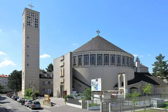 The image features a modern church building, characterized by its circular structure and a distinctive tall tower. It has a simplistic architectural style with a flat roof and features like a cross on top of the tower. Surrounding the church, there are a few trees and cars parked along the street, with clear blue skies providing a bright backdrop. The overall setting suggests an urban environment.

Image Credits: Wikimedia / C.Stadler/Bwag / CC BY-SA 4.0