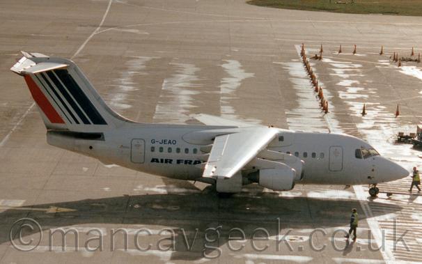 Side view of a high-winged, 4 engined jet airliner facing to the 
right but being pushed back to the left by an out-of-frame tug attached to the nose-wheel by a long pole.
The plane is mostly white, with dark blue "Air France" titles on the lower rear fuselage, with the black registration "G-JEAO" on the upper rear.
The tail has a series of dark blue diagonal stripes, thicker towards the front, with a single red stripe at the rear.
Grey concrete apron fills most of the rest of the frame, with a row of orange and white traffic cones in the background on the right, and a couple of people wearing hi-viz jackets wandering around.
Bright patches on the ground suggest that, while the sun bay be shining brightly, as suggested by the strong shadow this plane casts on the ground, but it was raining not too long ago.