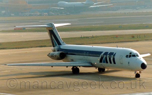 Side view of a twin engined jet airliner with the engines mounted on the sides of the rear fuselage, taxiing from left to right, following a white line on the taxiway.
The plane is mostly white, with dark blue "ATI" titles on the forward fuselage, with a lighter blue triangle filling the lower part of the letter "A", and a dark blue stripe running along the body, turning up into the tail where it repeats that letter "A".
There are black "DC-9" titles on the bare-metal engine pods on the sides of the rear fuselage.
Grey concrete apron fills the foreground, with large areas of green grass lining taxiways and a black runway in the background, with a grey and yellow terminal building stretching across the frame in the distance, with a very large, grey and blue jet airliner parked nose in in front of it.