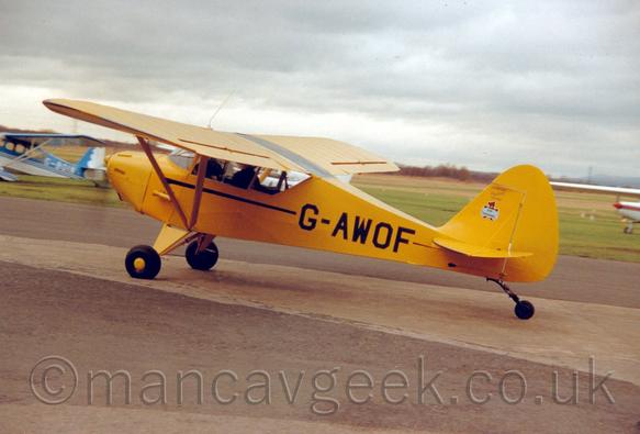 Side view of a high-winged, single engined light aircraft taxiing from right to left.
The plane is almost entirely yellow, with a thin black stripe running along the body, overlaid with the larger black registration "G-AWOF" on the rear fuselage.
A person with dark coloured clothes can be seen piloting the plane through the cockpit windows.
Grey concrete apron fills the foreground, with the flat grass airfield leading off to trees in the distance, under cloudy grey skies.