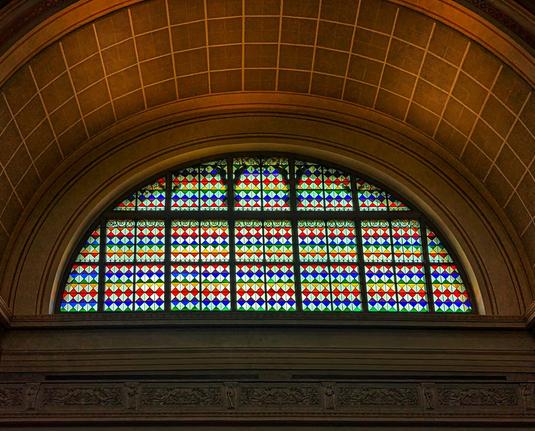 An ornate, arched stained-glass window dominates the centre of the frame, featuring a vibrant mosaic of red, blue, green, and yellow diamond and square shapes. It is set into a grand, light-brown archway with a textured, possibly coffered, ceiling above it. Below the window, intricate carved detailing is visible along the architectural elements. The lighting highlights the rich colours of the glass and the architectural grandeur of the structure.