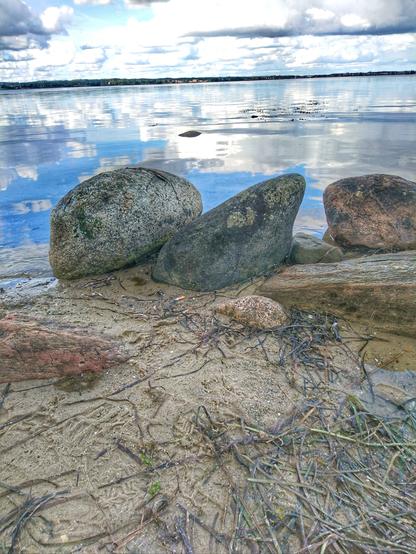 Am Ufer der Ostsee liegen Findlinge. Die Ostsee liegt still und flach da, fast wie ein Spiegel. Am Horizont ist das gegenüberliegende Ufer zu sehen.