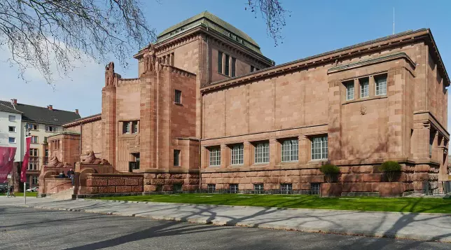 The image depicts a large building with a distinctive architectural style, characterized by its solid stone structure and prominent design elements. The building appears to have a symmetrical layout with various windows and ornamental details. It is likely set in a landscaped area, with some green grass and possibly trees in the vicinity. The overall impression suggests it is a significant public or cultural institution, such as a museum or gallery.

Image Credits: Wikimedia / Simsalabimbam / CC BY-SA 3.0