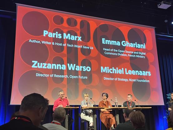 A photo showing four speakers on stage at a conference. From left to right: Michiel Leenaars, Zuzanna Watson, Emma Ghariani and Paris Marx. Above them there’s a tall large screen showing their names