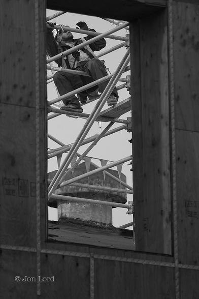 This Is A Black And White Street Photo In Portrait Format. Stretching Along The Left, Right And Lower Margins Are Panels Of Plywood Formed To Make Up An Opening Or Window Of About Two Metres Tall By A Little Over A Metre Wide: On A Construction Site. Framed Just Beyond The Window Is A Builder Sitting On A Lattice Of Scaffolding And Looking Upwards. He Has A White Hard Hat, Sunglasses And A Neatly Trimmed Dark Beard. He Is Wearing A Safety Harness Which Is Clipped To A Nearby Horizontal Strut. Behind And Below Our Contractor Is Part Of A Pitched Roof With A Large Cylindrical Vent Or Extractor Inserted Into The Roofs Surface.
The Location: The Embarcadero, San Francisco (2012).