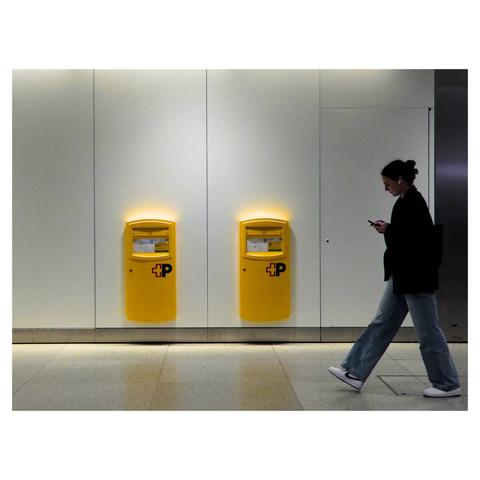 The image depicts an indoor railway station setting with a person walking past two yellow mail boxes mounted on a wall. The individual is dressed in casual attire, including a dark jacket, light blue jeans, and white sneakers, and appears to be looking at a smartphone. The background consists of a plain, light-colored wall and a tiled floor.