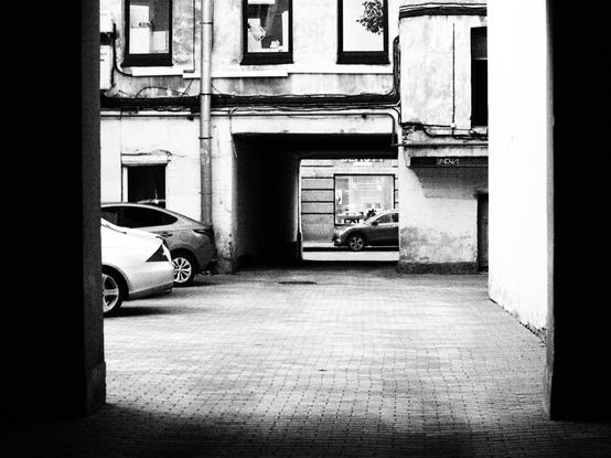 Black and white photo of a typical Saint-Petersburg courtyard in the center of the city. There are a two cars parked on the paving stones. And a passage to the street there the one car is riding somewhere.
The houses on the photo are entangled in wires.