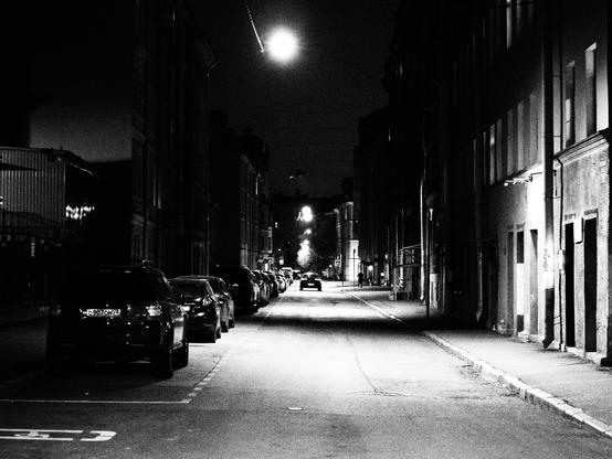The late evening photo of the typical Saint-Petersburg small street in the center of the city. The street is lit by lamps, hanged on the wire above the street. On the left there are a parking slots with a lot of cars in it. On the right there are living houses built in the end of XIX / start of XX centuries.
Photo is black and white.