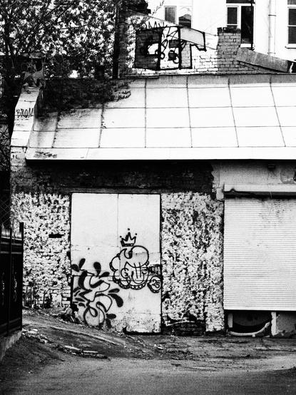 The  black and white photo of an old garage in the middle of Saint-Petersburg courtyards. The garage is built from bricks, has a tall doors, covered with unidentifiable graffiti tags and has a metal roof.