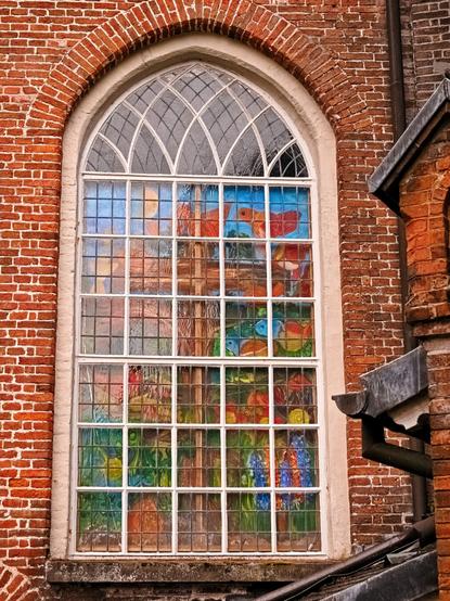 A church window in a red brick wall. The window is rounded at the top. It is painted with bright colours. You can see a brown cross surrounded by birds and other animals and plants. Rather naively drawn, and therefore I find it much nicer than the ‘artistically high-quality’ paintings that are usually found in/on churches.