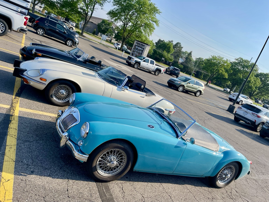 Photograph of three classic British sports cars in a parking lot with modern cars in the background. A blue MGA is in front with a white Jaguar E-Type behind and a black MGB behind that. The photograph tilts to the lower right.