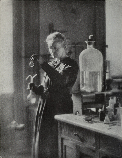 Madame Marie Sklodowska Curie Co-Discoverer of Radium Elected Honorary Fellow of The American Museum of Natural History, April 20, 1921.

Marie Curie stands alone, centrally positioned, wearing a long, dark, high-necked dress.

Her hair is tied back neatly, and she is shown in profile, with a calm, focused expression, absorbed in her scientific work.

She is holding a small flask in her left hand, examining it intently under the light.

In her right hand, she holds another laboratory implement, possibly a test tube or pipette, demonstrating active engagement in an experiment.

She is surrounded by lab equipment: on the counter are flasks, bottles, funnels, and other scientific glassware, suggesting a functional, hands-on workspace.

To her left (viewer’s right) is a large glass distillation or storage vessel, likely filled with liquid, reinforcing the laboratory context.

https://commons.wikimedia.org/wiki/Category:Marie_Curie#/media/File:Annual_report_of_the_trustees_of_the_American_Museum_of_Natural_History_for_the_year_(1921)_(19341550566).jpg