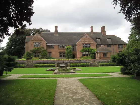 The image features a large, two-story brick house with a distinctive architectural style, surrounded by a well-maintained garden. The garden includes a central pathway leading to a rectangular area with plants or flowers and a landscaped lawn, contributing to an elegant outdoor setting. The sky is partly cloudy, and the surrounding trees add to the greenery and natural beauty of the scene.

Image Credits: Wikimedia / Bob Cantwell / CC BY-SA 2.0