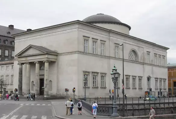 This image depicts a large, neoclassical building with a dome on top, situated at an intersection. The structure features a portico with columns, large windows, and a symmetrical design. There are people walking in the foreground, indicating it’s a public space. The overall appearance suggests it might be a cultural or governmental building.

Image Credits: Wikimedia / Ib Rasmussen / Public domain