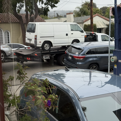 A view through our dining room window, over our neighbors' driveway and out to the street where a white circa-1990 Mitsubishi panel van is sitting on the back of a flatbed tow truck. The nose of our neighbors' silver Chevrolet Volt can be seen being towed off the back of the truck.