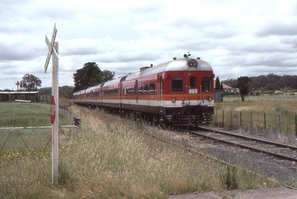 A 7-car DEB set diesel multiple unit train in Candy livery running on a single track line in a rural location approaches a level crossing marked with a set of crossbucks and a stop sign.