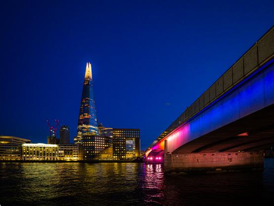 This is a stunning nighttime photograph of London's South Bank skyline taken from the River Thames. The image captures the dramatic contrast between the deep blue twilight sky and the illuminated cityscape below.

The Shard dominates the left side of the frame - London's iconic glass pyramid-shaped skyscraper rises majestically into the night sky, its upper floors glowing with warm golden light that creates a beacon-like effect against the dark blue backdrop.

The Bridge occupies the right portion of the image, stretching diagonally across the frame from bottom right toward the center. This appears to be London Bridge, illuminated with vibrant colored lighting that transitions from deep blue at the top to brilliant magenta and pink at the water level. The bridge's concrete structure shows clearly defined architectural details and support pillars.

The Water in the foreground is the River Thames, appearing as a dark, rippling surface that perfectly mirrors the colorful bridge lighting a
