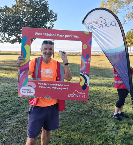 Orange shirted man with finish  tokens in hand under the Wes Mitchell parkrun sign with Tin Can bay estuary behind