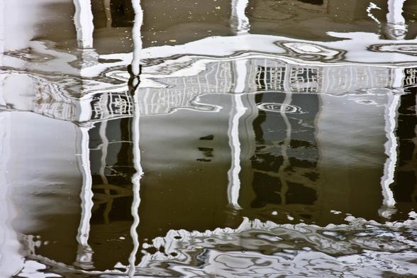 A reflection of the front of a stately, white Hague villa in a canal. You only see a terrace and some windows in the photo, upside down. The image in the water has become black and white. The poles of the terrace and the styles of the windows are a bit crooked by the wind. The windows look like dark holes. The whole thing gets the atmosphere of a lost place.