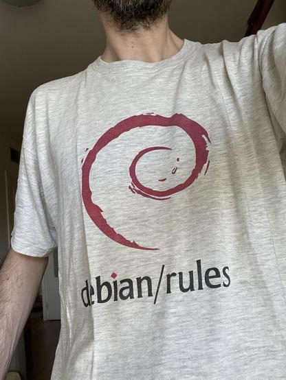 Yo llevando una camiseta gris con el logotipo de Debian, una espiral roja pintada con brocha, sobre el texto “debian/rules”