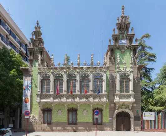 The image depicts a decorative building with a mix of architectural styles, prominently featuring ornate facades with intricate details. The building is adorned with sculptures and has a distinct color palette, primarily featuring green and beige. Flags are visible on the front, and the setting appears to be a street with some trees nearby. The structure reflects a historical or cultural significance, likely located in an urban environment.

Image Credits: Wikimedia / Diego Delso / CC BY-SA 4.0