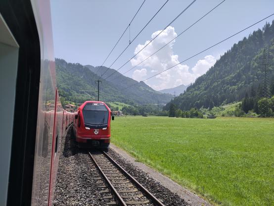 Foto. Ein roter Zug in Berglandschaft, aus dem Fenster eines weiteren Zuges betrachtet.