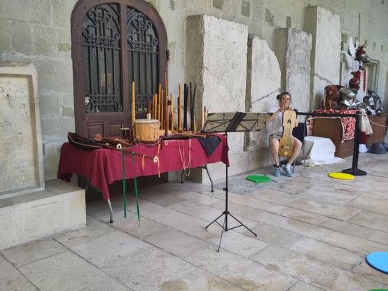 A table covered in wooden instruments (pipes, drums, strings), with someone tuning a 5-string instrument with a modern tuner.
