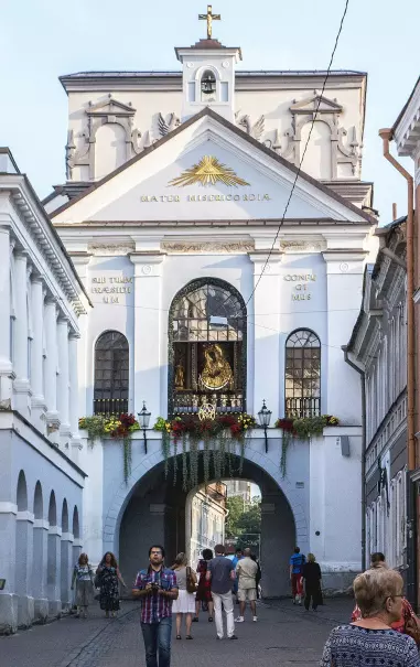 The image depicts an architectural structure, likely a historical or cultural landmark, featuring a white facade with ornate details and a cross at the top. There are people walking through a street and passing under an archway that is decorated with flowers. The setting appears to be a vibrant urban area, possibly in a European city. The overall atmosphere suggests a combination of historical significance and everyday life, with pedestrians engaging in typical activities.

Image Credits: Wikimedia / Jerzy Strzelecki / CC BY-SA 3.0