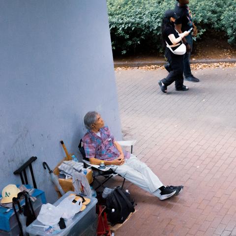 Elderly person sitting in a folding chair against a wall on a paved area. Surrounding them are personal belongings including a suitcase, a container, an umbrella, and a backpack. Two people walk on a path bordered by greenery in the background.
