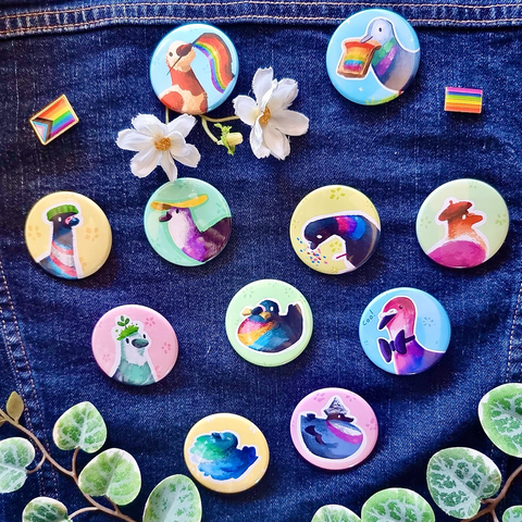 Picture of a set of 11 different button badges designs pinned to a jean jacket. Each badge design represents different LGBTQ flag colors displayed through pigeons in their colouring and accessories.