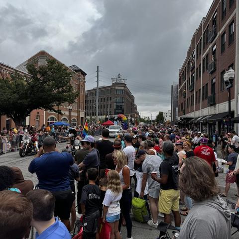 A wide street filled with large crowds attending a pride parade. Spectators of all ages line the sidewalks, facing toward floats and participants farther down the street. Several people hold rainbow flags or wear colorful outfits. Urban buildings frame both sides of the street under a cloudy sky, with the “Bottleworks District” sign visible in the distance.