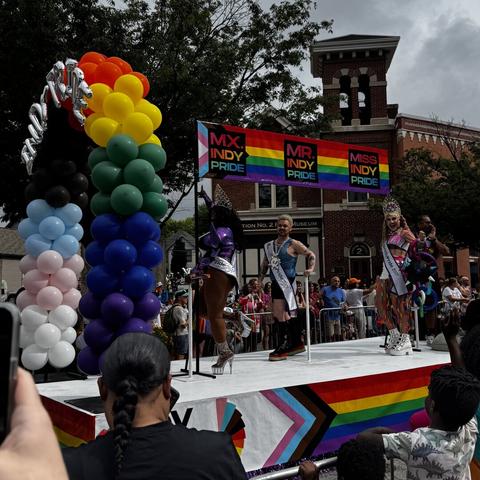 A pride parade float with a rainbow balloon arch and a rainbow banner labeled “Mx. Indy Pride,” “Mr. Indy Pride,” and “Miss Indy Pride” with people wearing sashes with the same labels standing underneath. People watch and take photos from the crowd along the street, with trees and a brick building in the background.