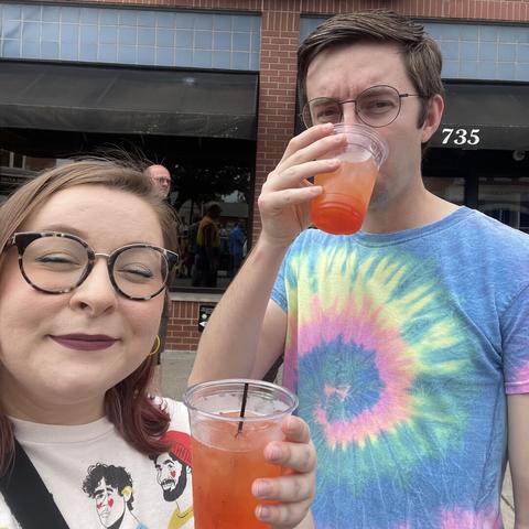 A smiling couple stand outside of a brick building wearing colorful clothing. They are both holding a red/orange drink in clear plastic cups.