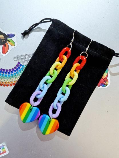 Grey, metallic surface (closed laptop) with rainbow stickers. On it lies a black sachet, and on that are long earrings in rainbow colours: a colourful thick chain with a rainbow heart at the end.