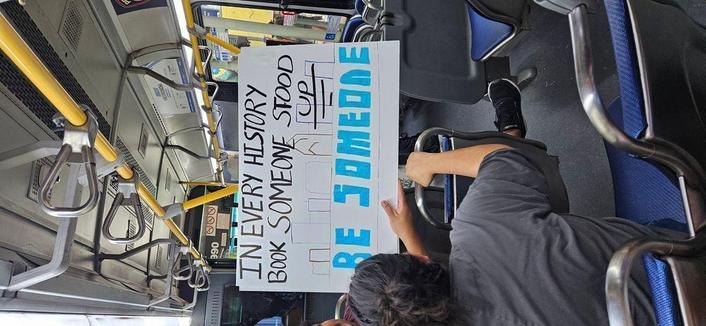 Sign on a bus headed to protest that reads "IN EVERY HISTORY BOOK SOMEONE STOOD UP. BE SOMEONE." "Be someone" is written in block letters in the color blue, which mimics local graffiti above a bridge over I-45N southbound just outside downtown Houston.