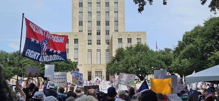 Photo of protest crowd in front of Houston City Hall with lots of signs visible. From left to right: A red and blue flag with a fist covered in the American flag states "RESIST FASCISM. FIGHT OLIGARCHY;" "PATRIOTS PROTEST TYRANNY;" "IMMIGRANTS DON'T SCARE ME. KINGS DO!;" "NO KINGS JUST AMERICANS;" "REPUBLICANS NOW OK WITH KINK AT PRIDE" with a coiled snake saying "Tread on me, Daddy."