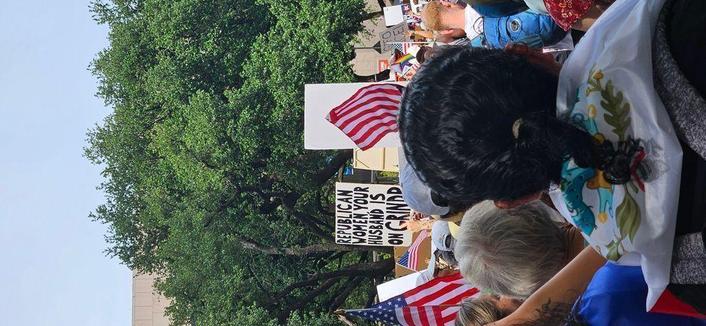 In the foreground, back of people's heads at a protest. In the middle, a sign reads, "REPUBLICAN WOMEN YOUR HUSBAND IN ON GRINDR." In the background, trees below a blue sky.